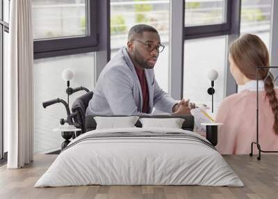 Disabled male boss has dark skin, dressed in formal cothes, analyzes budget receipts and outlays together with female assistant, can`t understand something in graphics, sit over loft office interior Wall mural